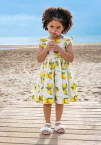 Robe blanche avec imprimé citron jaune, manches courtes à volants et taille froncée. L'enfant tient des fleurs, portant des sandales blanches avec des accents volantés.