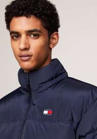 Giacca puffer blu navy con colletto alto e chiusura con zip, arricchita da un piccolo logo patch di Tommy Jeans sul petto. Texture liscia e lucida.