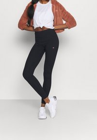 Leggings negras con una cinturilla alta, que presentan un pequeño logo en la cadera. Combinados con una camiseta blanca y una chaqueta fuzzy naranja.