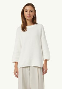 White, ribbed knit sweater with a round neck, dropped shoulders, and three-quarter sleeves. Worn over light beige, pleated trousers.