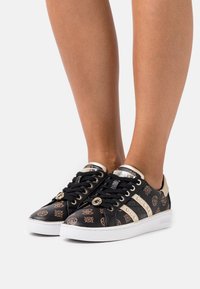 Black sneakers with gold accents, patterned detailing, glossy material, round laces, and a white sole. "Guess" logo on the side panel.