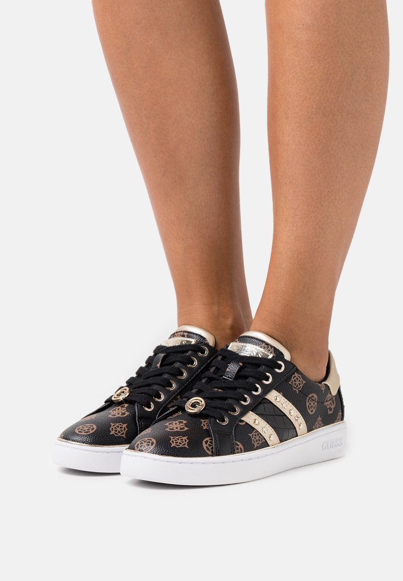 Black sneakers with gold accents, patterned detailing, glossy material, round laces, and a white sole. "Guess" logo on the side panel.