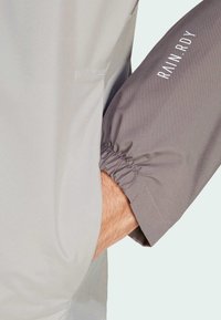 Lekka, szara kurtka przeciwdeszczowa z elastycznymi mankietami, z kontrastowym ciemnoszarym rękawem i nadrukiem "RAIN.RDY" na biało. Gładka faktura.