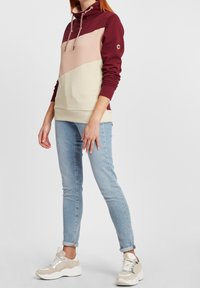 Personne portant un sweat à capuche color-block en bordeaux, rose et crème, un jean skinny bleu clair et des baskets beige et blanc, debout devant un fond blanc.