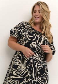 Blonde Frau in einem schwarz-weißen Kleid mit abstraktem Druck, die das Taillenband anpasst, mit geschlossenen Augen und sanftem Lächeln vor einem weißen Hintergrund.