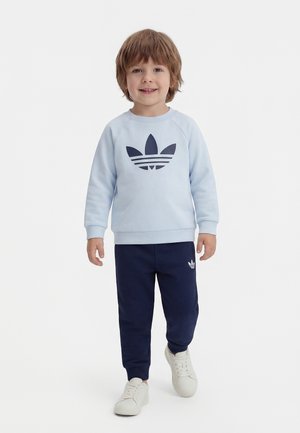Young child walking forward wearing light blue sweatshirt and navy pants with Adidas logos, white sneakers, smiling, neutral background.