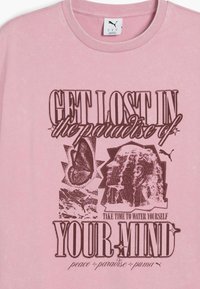 T-shirt di cotone rosa chiaro con grafiche stampate di una farfalla, un viso e testo in marrone. Collo rotondo e maniche corte.