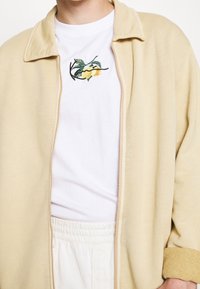 Krämfärgad zip-up jacka med mjuk textur, kombinerad med en vit t-shirt med färgglad citronbrodyr. Lätta, avslappnade byxor med lös passform.