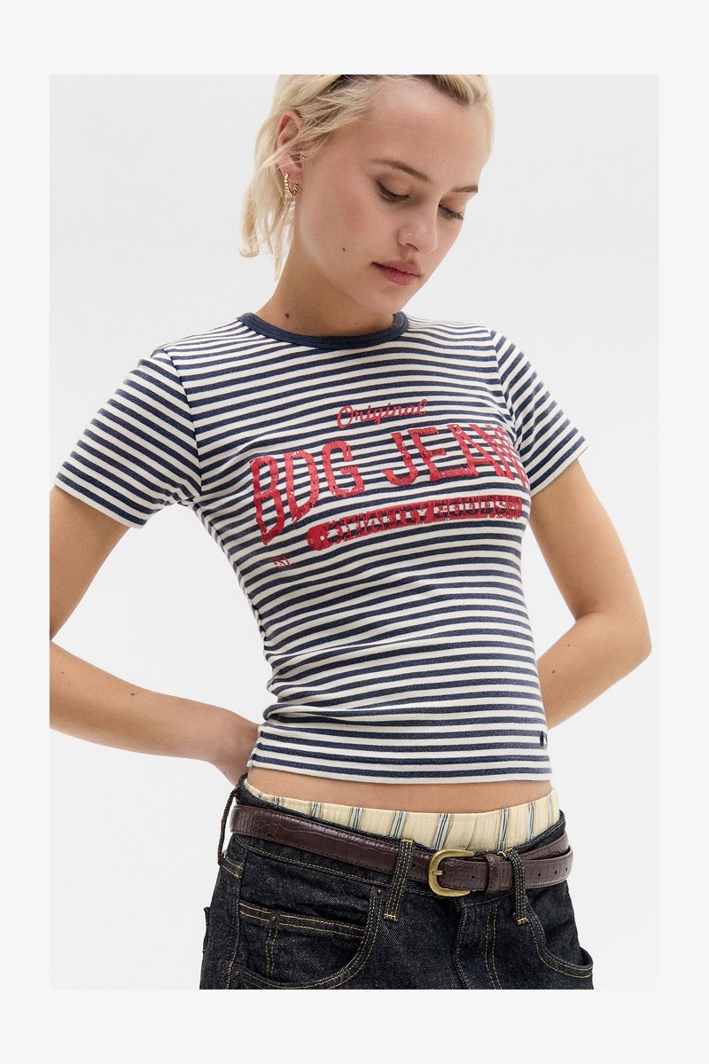 Nő tengerészkék és fehér csíkos BDG Jeans pólóban, sötét farmerben barna övvel, állva, kezét a csípőjén tartva, lefelé néz.