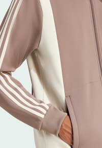 Giacca con zip anteriore in malva e crema, con tessuto testurizzato, tasche laterali e tre strisce bianche sulle maniche.