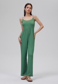 Jumpsuit vert à côtes avec des bretelles fines. Présente un corsage ajusté, une taille élastique et une conception à jambe large. Texture lisse, style décontracté.
