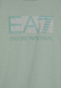 Světle zelené tričko s vyvýšeným logem "EA7" a geometrickými vzory, s textem "EMPORIO ARMANI" v mírně tmavší barvě.