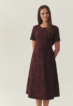 Kobieta ubrana w ciemnofioletową sukienkę do kolan z paskiem, zdobioną kwiatowym wzorem, stoi z rękami z tyłu na prostym tle.