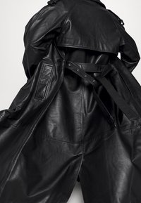 Černý kožený trench coat s páskem v pase, širokými klopami a volným střihem. Obsahuje boční kapsy a texturovaný kožený povrch.
