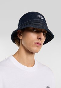 Cappello bucket in denim blu scuro con logo del marchio sul davanti, dotato di una ampia visiera inclinata verso il basso e dettagli di cucitura visibili.