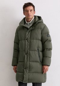 Donkergroene gewatteerde jas met capuchon, voorzien van een ritssluiting aan de voorkant, zijzakken en een grijze, gestructureerde trui eronder.