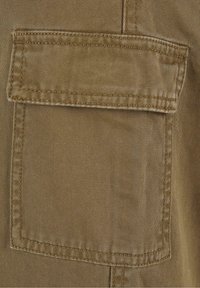Gros plan d'une poche en tissu kaki avec rabat, détaillée par une double surpiqûre le long des bords et des lignes de couture.