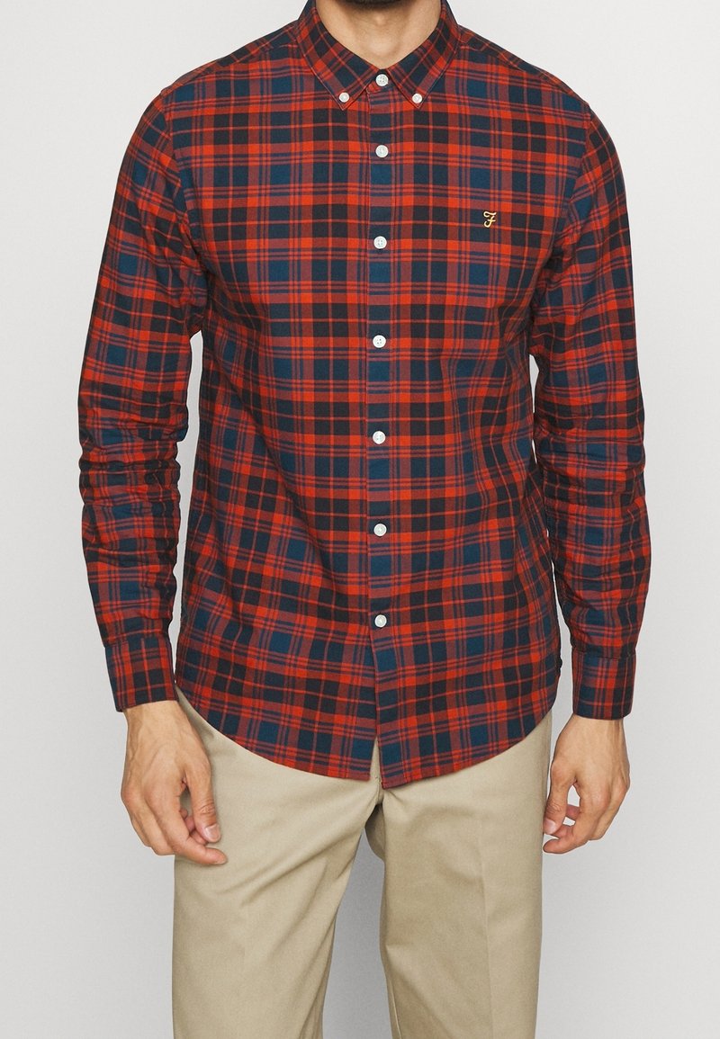 Homme portant une chemise à carreaux rouge et bleu marine boutonnée et un pantalon beige, debout les bras détendus le long du corps.