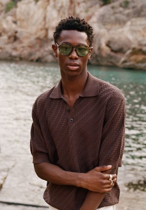 Fiatal férfi barna texturált galléros pólót és napszemüveget visel, karját keresztbe téve áll a sziklás vízpart mellett, a kamerába nézve.