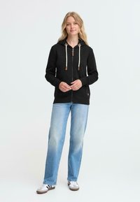 Crna zip-up jakna s kremastim vezicama i smeškastim detaljima, uparena s svijetloplavim trapericama ravnog kroja i sivoj niskoprofilnoj obući.