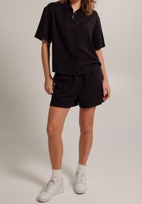 Černá lněná košile s krátkým rukávem a knoflíkovým zapínáním, s kapsou a ladícími šortkami, v uvolněném střihu, doplněná o bílé tenisky a ponožky.