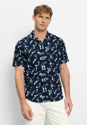 Mann trägt ein marineblaues Kurzarmhemd mit weißen Tennis-Spieler- und Tennisschläger-Motiven, kombiniert mit weißen Hosen, steht vor einem schlichten Hintergrund.