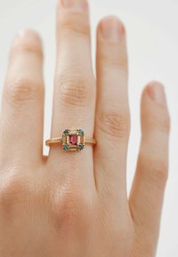 Anillo de oro con una piedra central en corte esmeralda de color rosa, rodeada de acentos azules, con una banda texturizada. Se lleva en una mano con textura de piel visible.