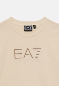 Bežs T-krekls ar apaļu kaklu, ar reljefu izsistiem "EA7" un "Emporio Armani" logotipiem kontrastējošās krāsās. Izgatavots no mīksta kokvilnas materiāla.