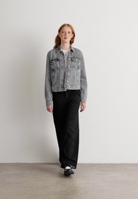 Grijze denim jas met knoopsluiting en twee voorkruipzakken, gecombineerd met zwarte wijde broek en zwarte schoenen, staand op een neutrale achtergrond.