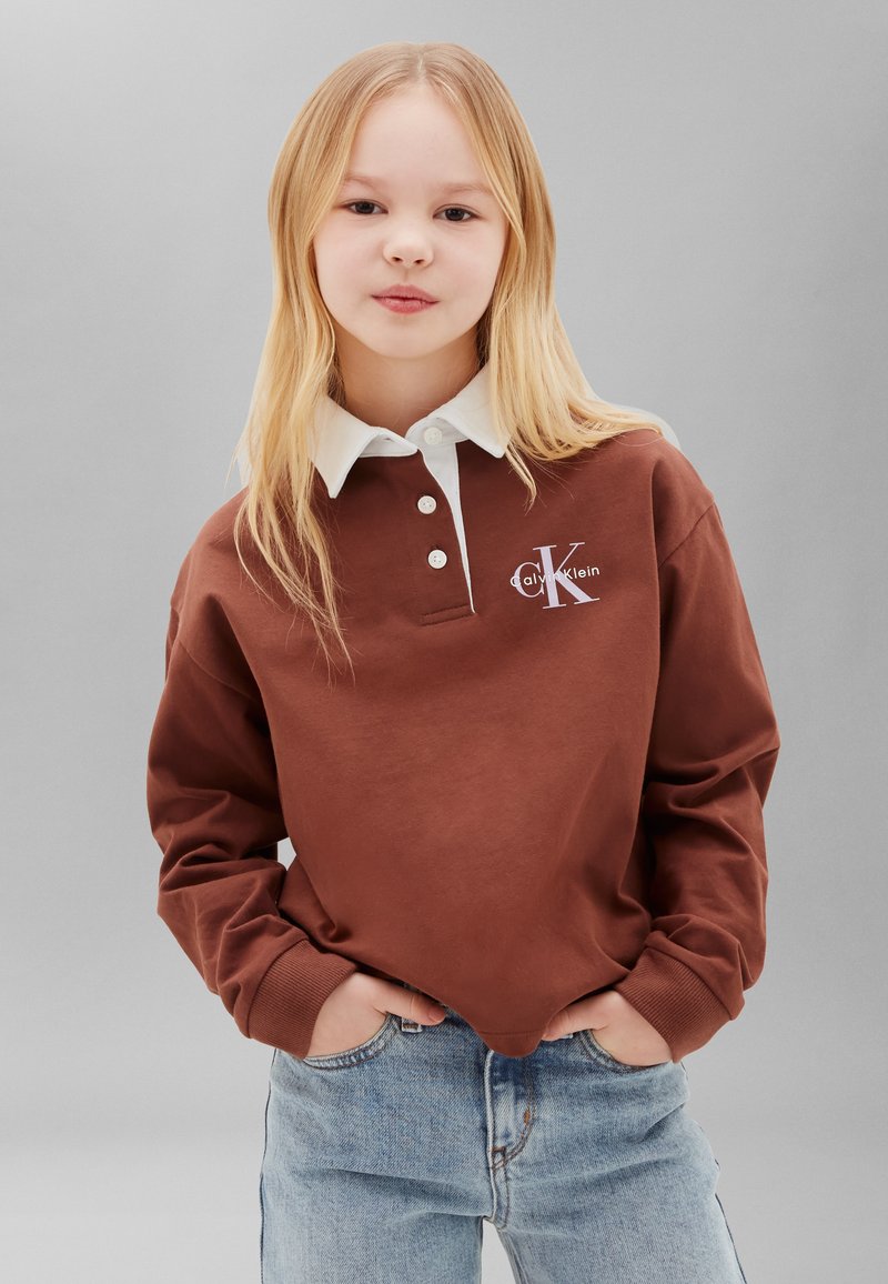 Ξανθό παιδί που φοράει ένα καφέ sweatshirt Calvin Klein με λευκό γιακά και ανοιχτό μπλε τζιν, στέκεται με τα χέρια στις τσέπες.