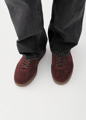 Bordeauxrote Wildleder-Sneaker mit braunen Gummisohlen, in einem flachen Schnitt mit flachen Schnürsenkeln, kombiniert mit schwarzen Denim-Hosen.