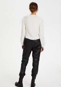 Blusa bianca con arricciature e maniche a sbuffo; abbinata a pantaloni in pelle nera e stivali alla caviglia, che evidenziano il tessuto strutturato e la vestibilità sartoriale.