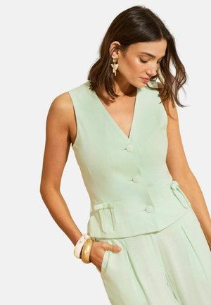 Olkapäille ylettyvät ruskeat hiukset omaava nainen pukeutuu hihattomaan mintunvihreään napitettuun liiviin ja siihen sopiviin housuihin, joita koristavat kullan- ja valkoisenväriset rannekorut.