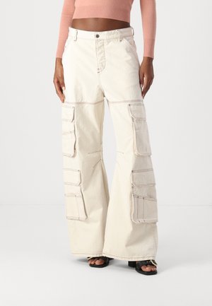 Persoon die roomkleurige cargo-broek met hoge taille en meerdere grote zakken draagt, gecombineerd met een kort perzikkleurig longsleeve topje, staand op een witte vloer.