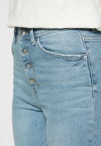 Jeans de ganga azul claro com cinco botões de metal à frente, desgaste subtil e bolsos normais nas laterais e atrás.