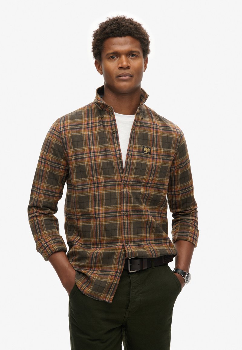 Camisa de cuadros marrones con acentos en amarillo, rojo y verde, con cuello abotonado y bolsillo en el pecho, combinada con pantalones verdes oscuros.