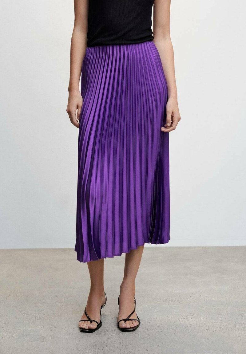 Femme portant une jupe midi plissée violette, un top noir sans manches et des talons ouverts à brides noirs, debout sur un sol en béton.