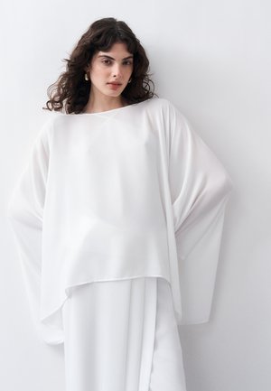 Γυναίκα με κυματιστά μαλλιά που φοράει ένα άνετο, ρέον λευκό τοπ και φούστα, στέκεται μπροστά σε ένα απλό λευκό φόντο.