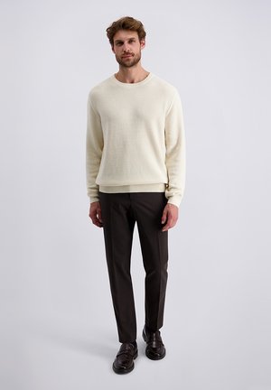 Un pull en maille texturée beige clair à manches longues, associé à un pantalon sur mesure marron foncé et des chaussures noires à enfiler.