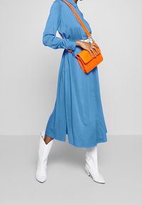 Robe bleue à manches longues avec une taille ceinturée, associée à un sac bandoulière orange texturé et des bottines blanches à talons. Tissu lisse et couleurs vives.