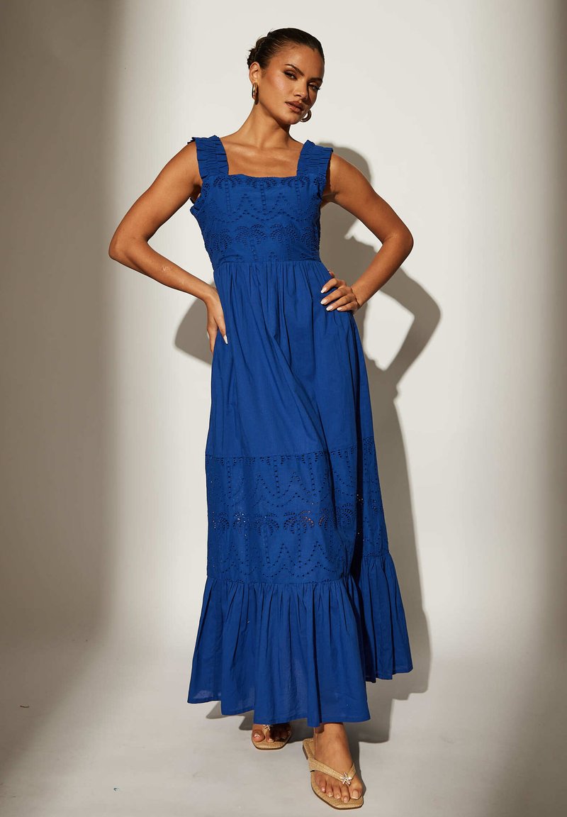 Vestido largo de algodón azul con diseño de capas, tirantes con volantes y patrones cortados. Parte inferior acampanada y combinado con sandalias planas color marrón claro.