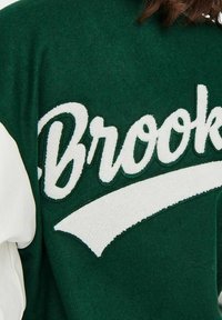 Zielony sweter z okrągłym dekoltem z białym nadrukiem „Brooks”. Charakteryzuje się miękką fakturą oraz kontrastowymi białymi akcentami na rękawach.