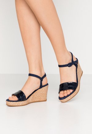 Navy wedge sandals feature a glossy croc-embossed strap, adjustable ankle buckle, and a woven jute platform for texture and support.