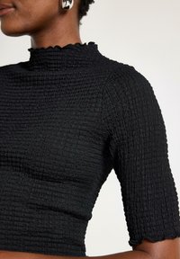 Sort smocked top med en høj, bølget krave, korte ærmer og struktureret stof. Detaljer inkluderer et rynket design og en tætsiddende silhuet.