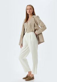 Blazer beige con bottoni dorati sopra un top cropped beige chiaro e pantaloni bianchi a pieghe. Mocassini marroni con texture completano l'outfit.