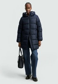 Tumši zils pūkains mētelis ar kapuci, priekšā rāvējslēdzējs, taisnstūrveida siluets un horizontālas šuves. Valkāts kopā ar platu džinsu biksēm un somiņu.