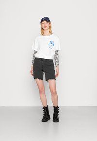 T-shirt branca com gráfico de mão azul, sobreposto com uma camisa de manga longa às riscas preta e branca, calções de ganga pretos e botas de combate pretas.
