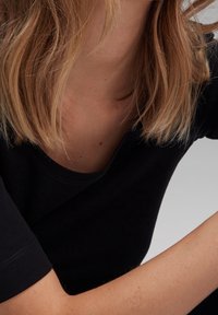 Blonde woman in a black top with arm bent, showing her neck, shoulder-length hair, and upper arm against a light background.