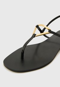 Sandalo piatto nero con suola liscia, caratterizzato da un accento di hardware triangolare dorato e sottili cinturini neri con cuciture bianche.