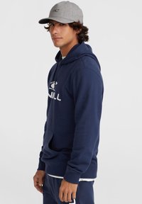 Navy blue hooded sweatshirt with a front pocket and white logo. Worn with a gray cap, featuring a curved brim and a subtle logo.